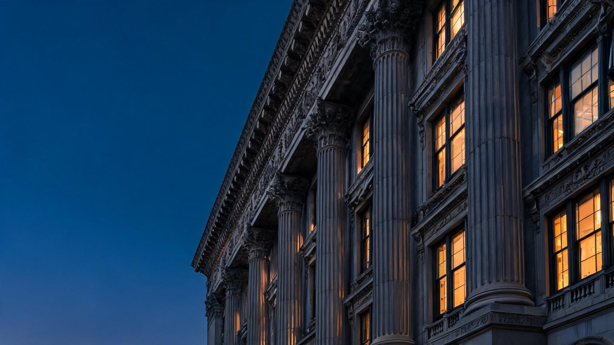 Stone facade of a classical building with columns and lit windows at dusk.